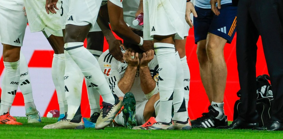 a group of soccer players huddle around a player who is laying on the ground