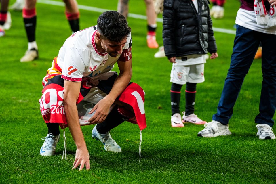 A Sevilla FC player kneels on the field after his last professional game, draped in a commemorative scarf.