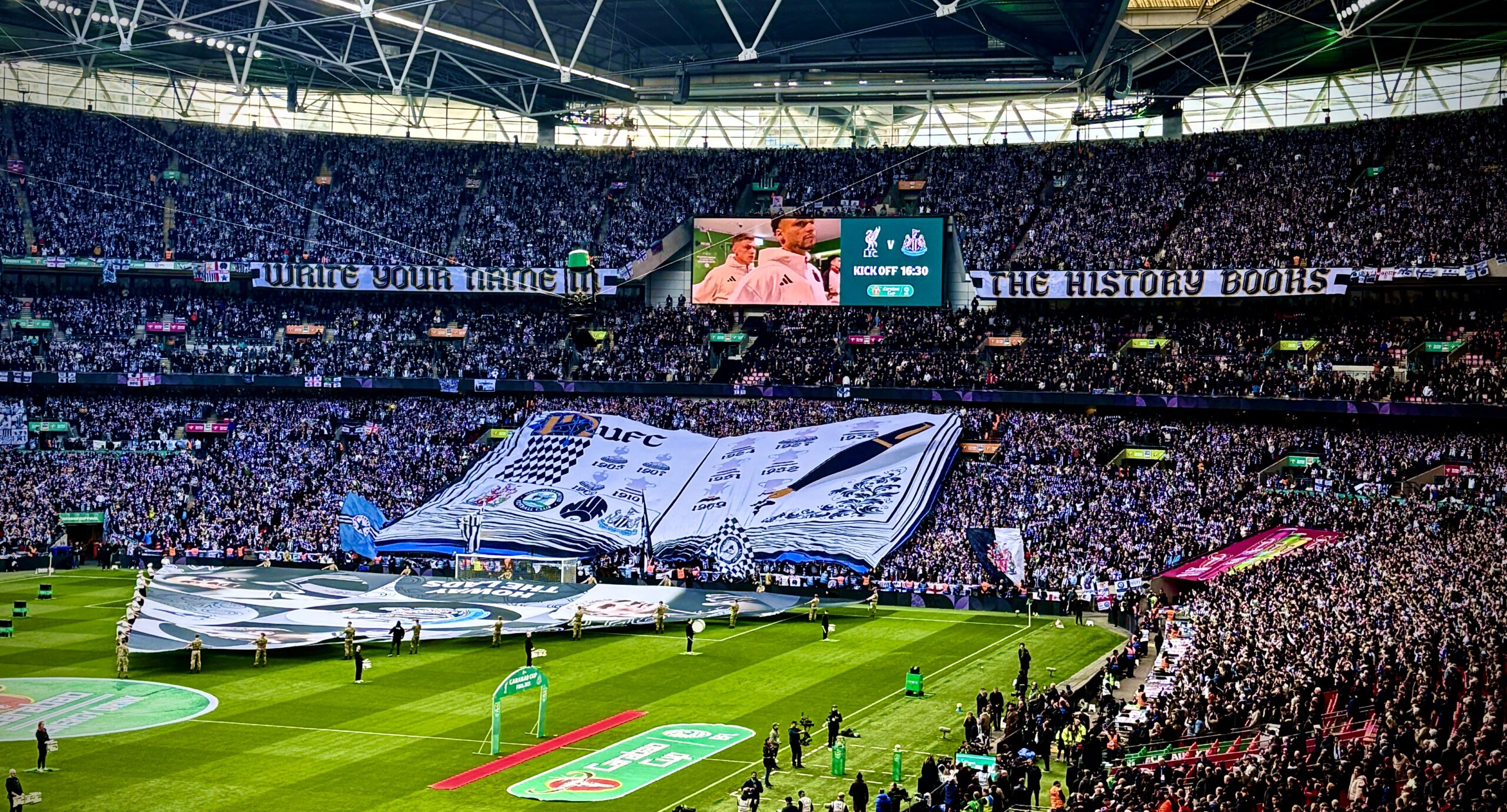 Newcastle United fans unveil a TIFO display curated by supporter group Wor Flags that says 'Write Your Name In The History Books' and features a quill and book. Image taken by journalist Matthew Ketchell